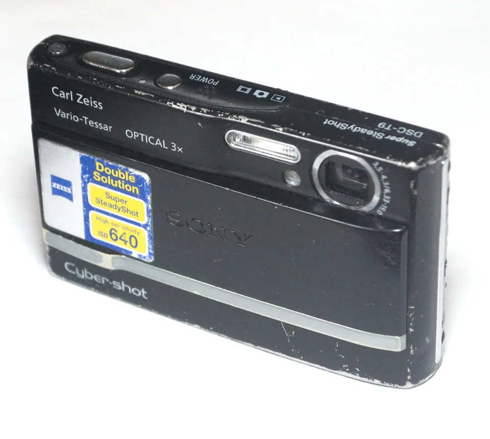 A well-worn black Sony Cyber-shot DSC-T9, front view, with its sliding door open.